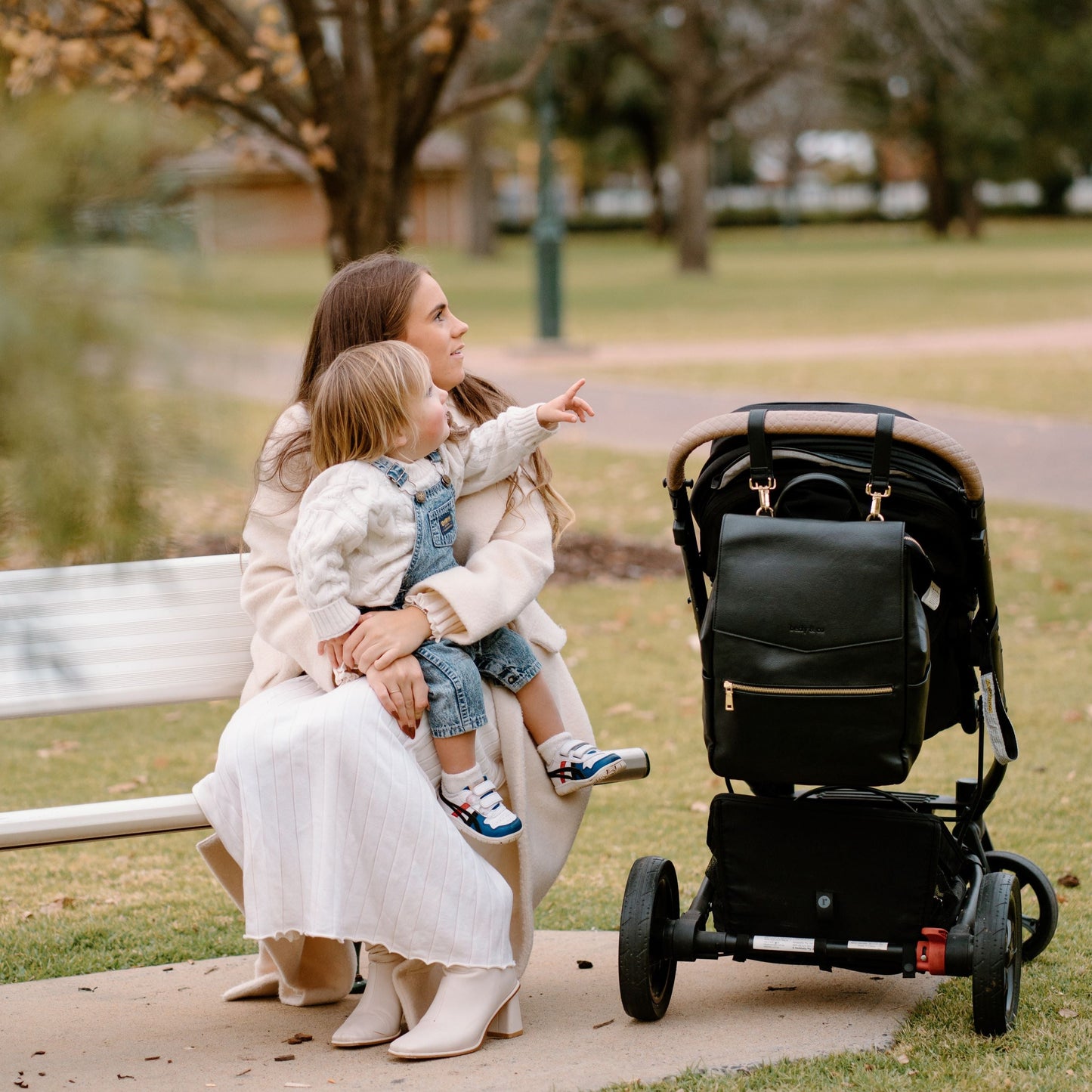 Parent and toddler in the park with Florence black nappy bag hanging off stroller with pram straps 