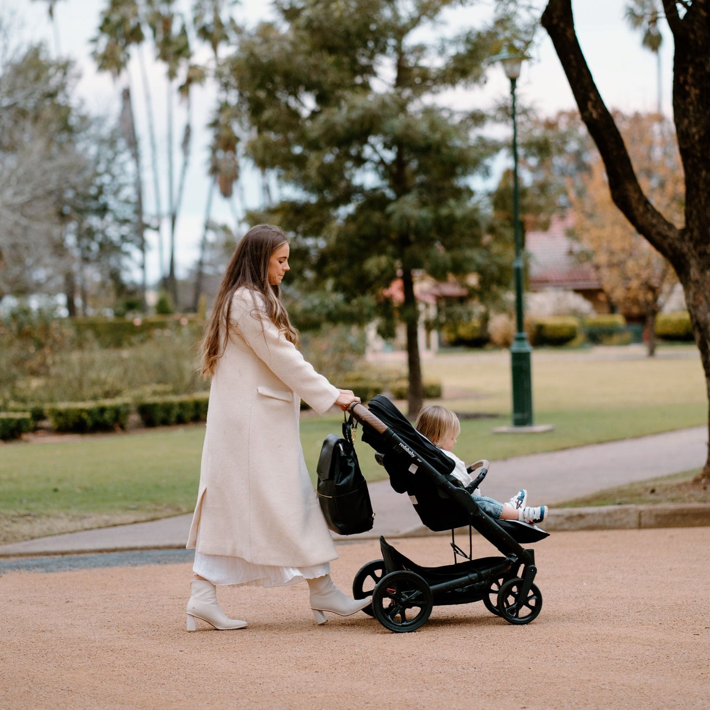 Parent and toddler in the park with Florence black nappy bag hanging off stroller with pram straps 